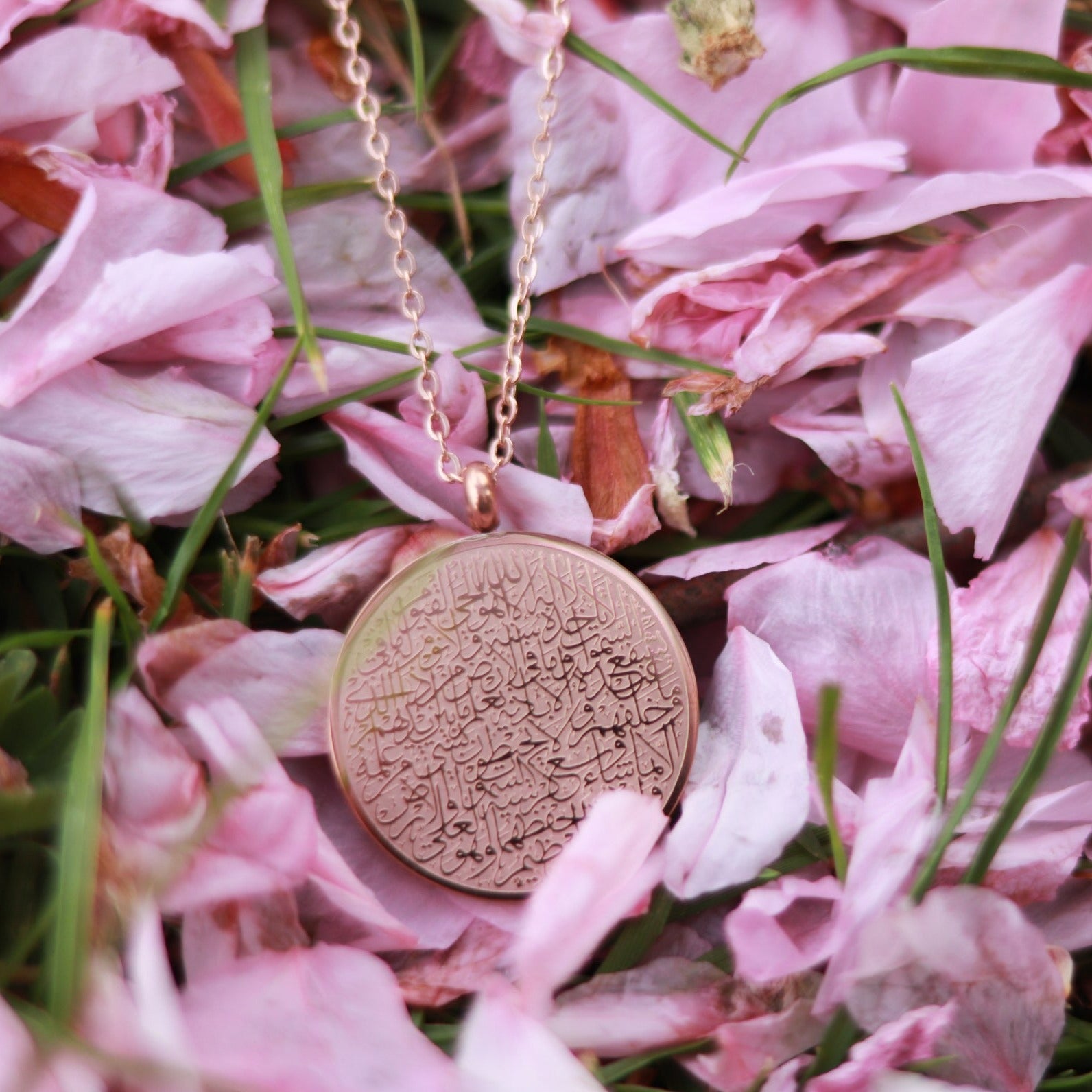 Rosé Halskette mit rundem Anhänger auf dem in arabischer Schrift und feinster Kalligrafie Ayatul Kursi, der Thronvers eingraviert ist. Die Halskette liegt auf einer mit Kirschblütenblättern beschmückten Wiese