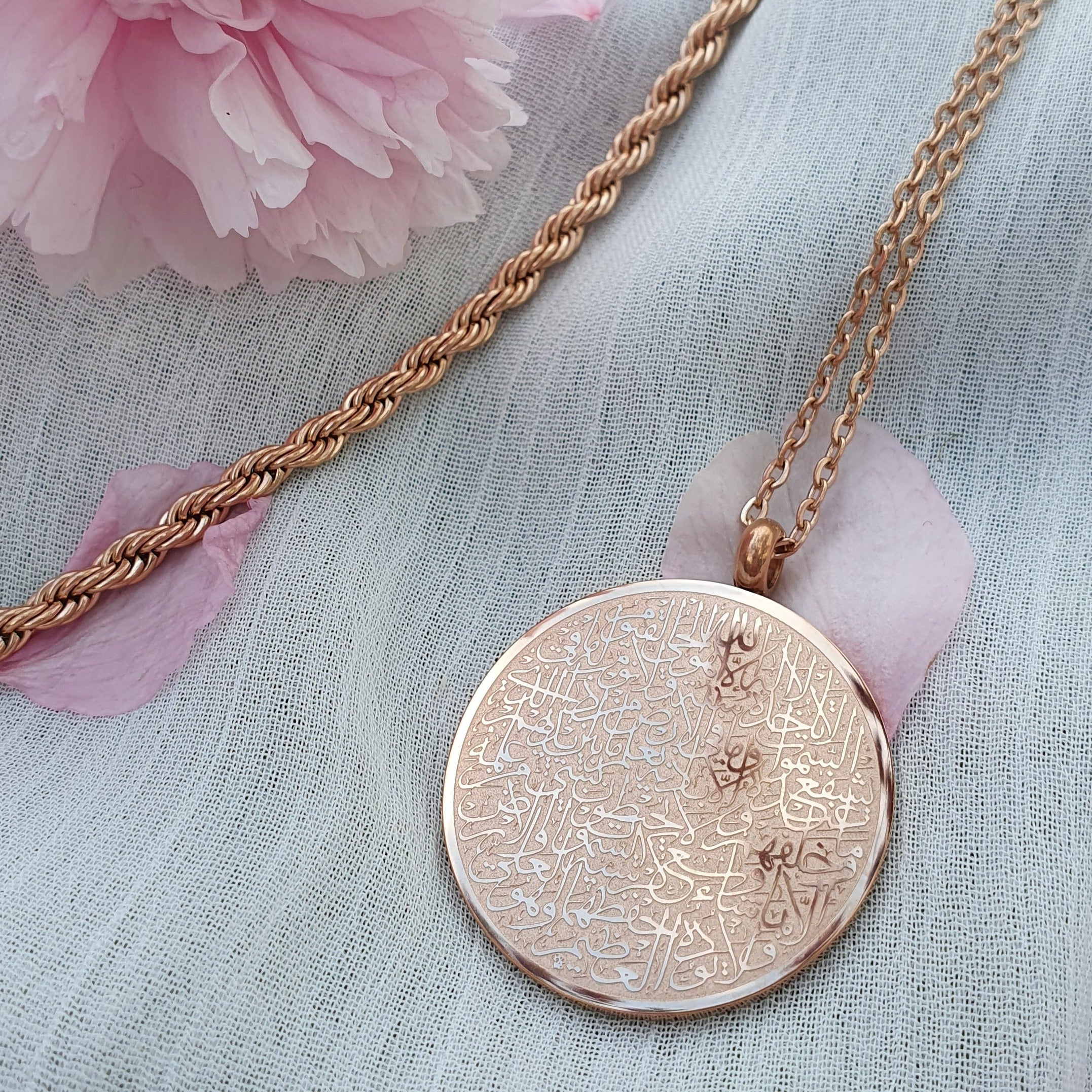 Goldene 18 Karat vergoldete Halskette mit rundem Anhänger auf dem in arabischer Schrift und feinster Kalligrafie Ayatul Kursi, der Thronvers eingraviert ist. Die Halskette liegt auf einem weißen Untergrund, auf dem Kirschblütenblätter liegen