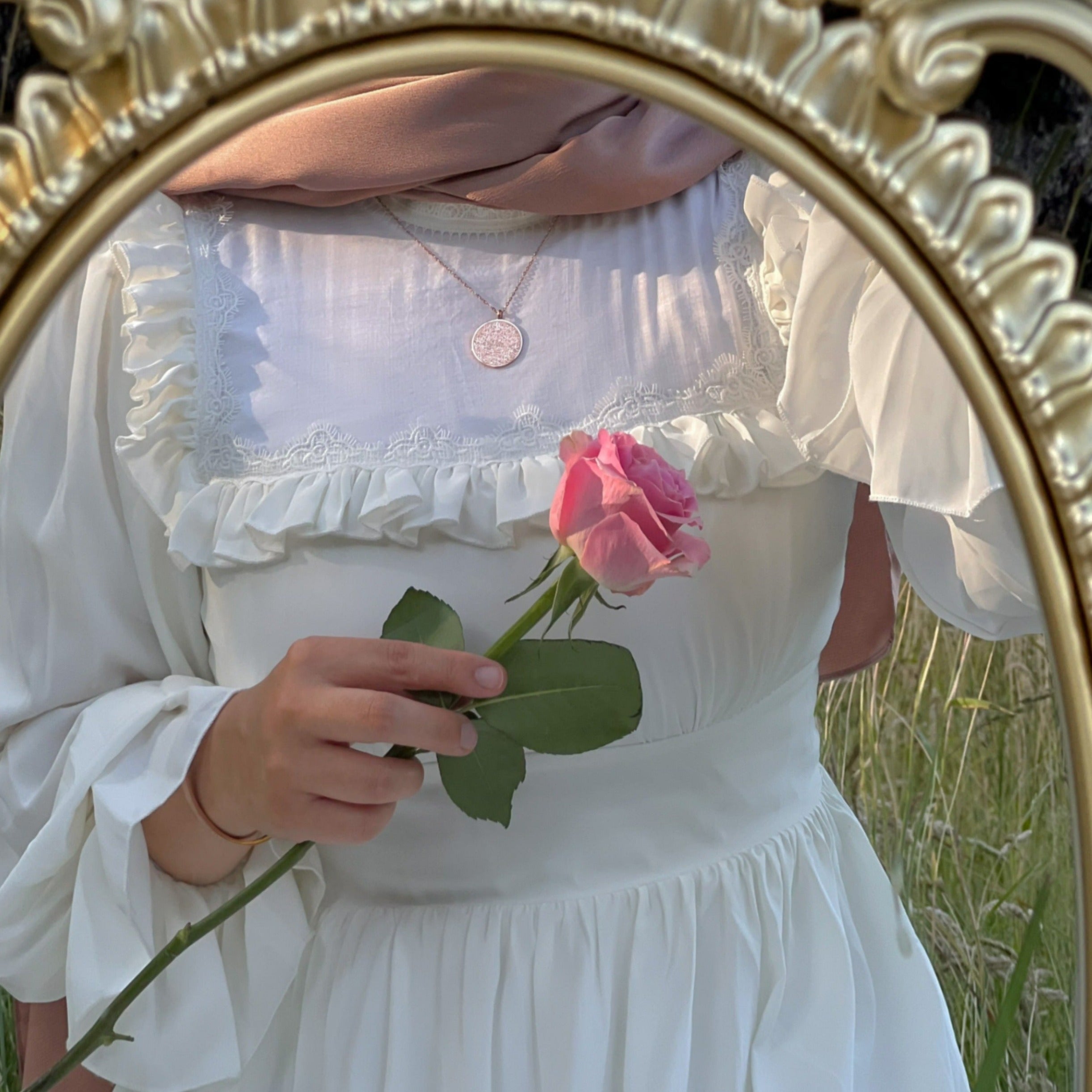 EIn goldener pompöser Spiegel, in dem sich ein Mädchen spiegelt. Sie trägt ein weißes Kleid mit Rüschen und ein braunes glänzendes Kopftuch. In ihrer Hand hält sie eine rosafarbene Rose. An ihrem Hals baumelt eine wunderschöne Halskette mit rundem Anhänger, auf dem der Thronvers, Ayatul Kursi, eingraviert ist.
