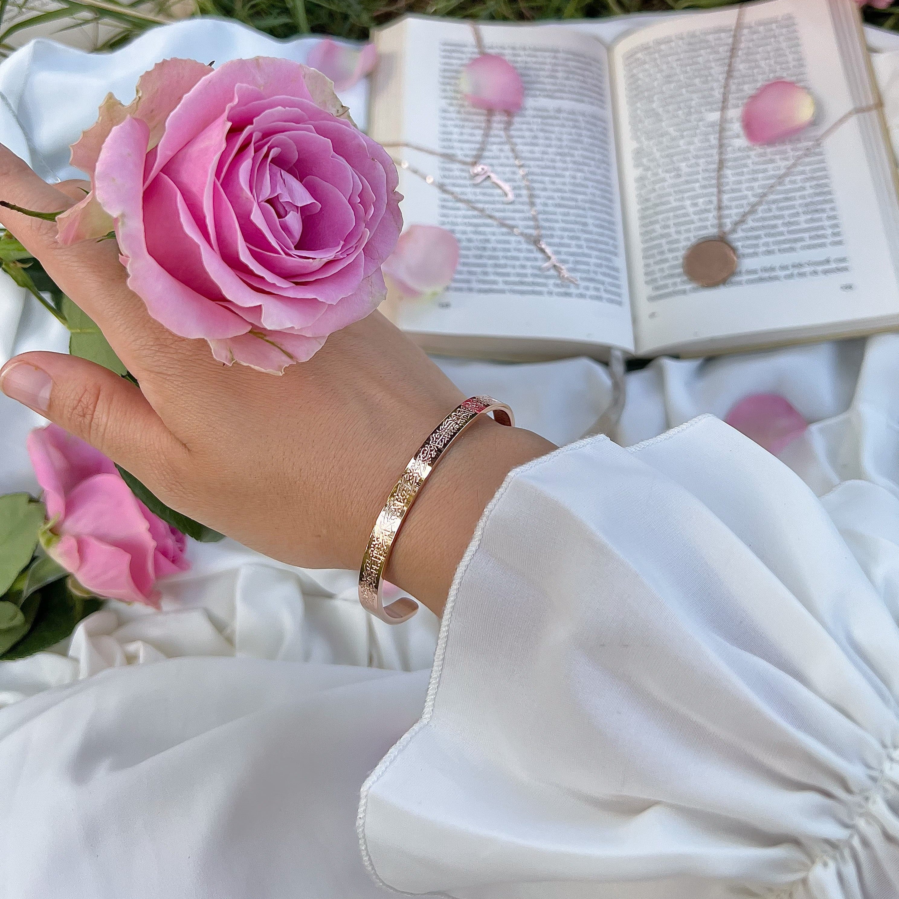 Im Vordergrund: Eine Hand, die eine wunderschöne rosafarbene Rose hält und am Handgelenk einen roséfarben Armreif mit Ayatul Kursi darauf trägt. Die Trägerin trägt ein weißes Kleid mit Rüschendetails am Handgelenk. Im Hintergrund liegt ein aufgeschlagenes Buch mit Rosenblüten darauf und weiteren Schmuckstücken von Hilaling